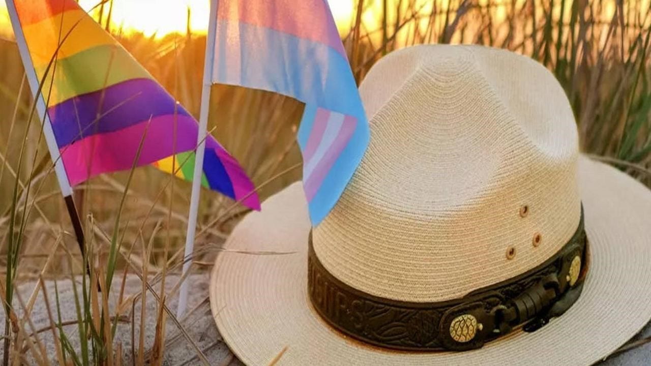 A park rangers flat hat sits atop a sandy dune with an LGBTQ pride flag and Transgender pride flag next to it.