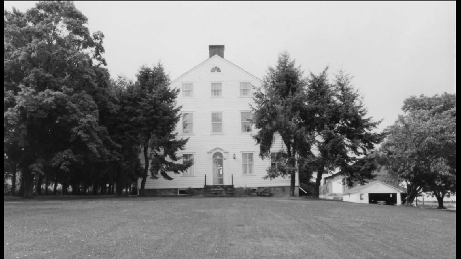 A black and white photograph of a large three story home with white siding and several pine trees in the front yard.