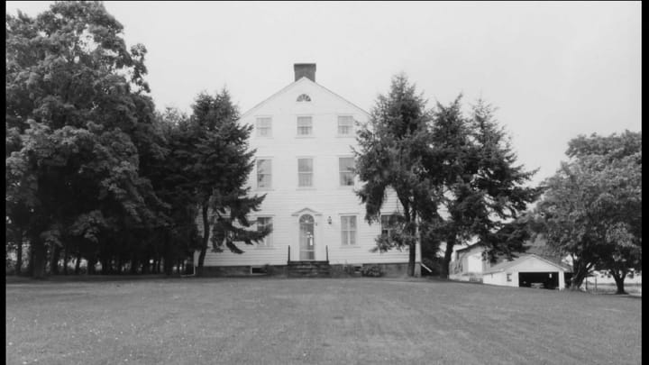 A black and white photograph of a large three story home with white siding and several pine trees in the front yard.